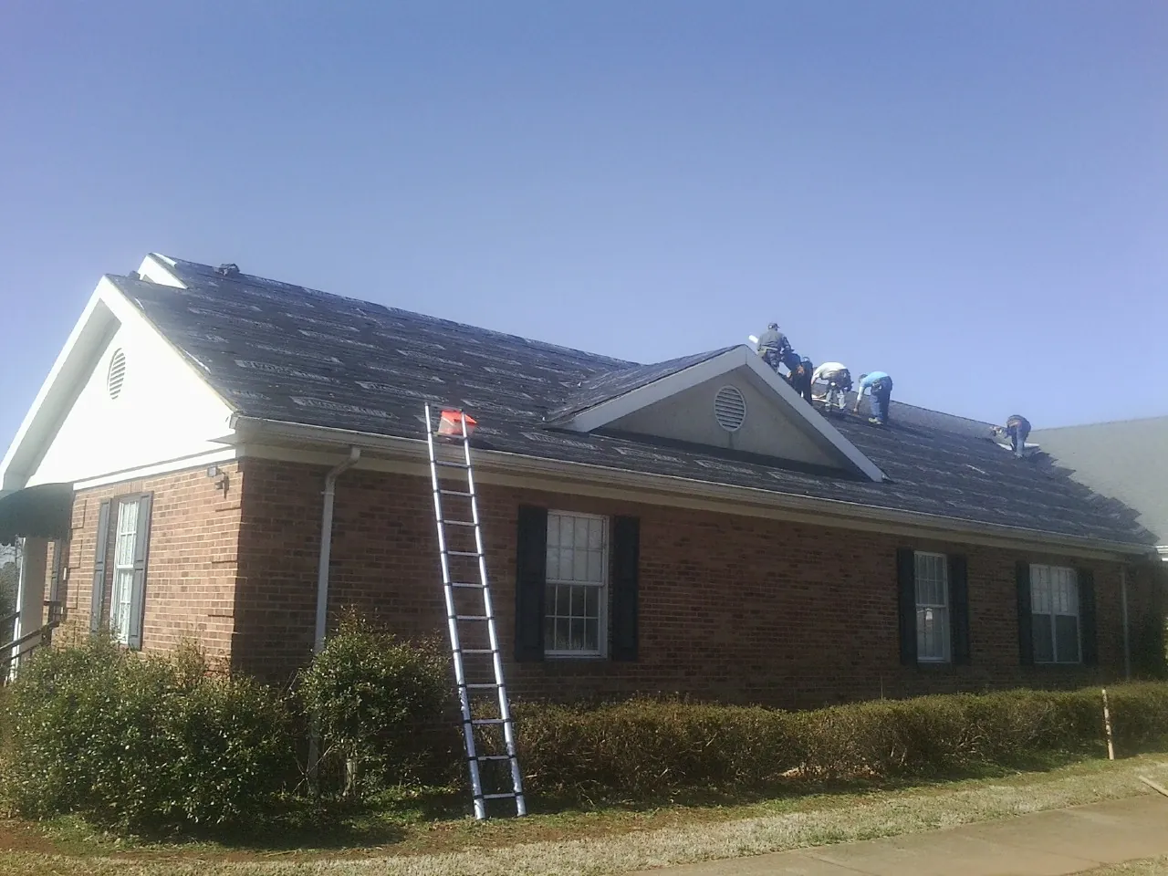 Crew tearing shingles off a brick ranch roof with a ladder set — Metal Roof Repair work by our crew in Huntertown