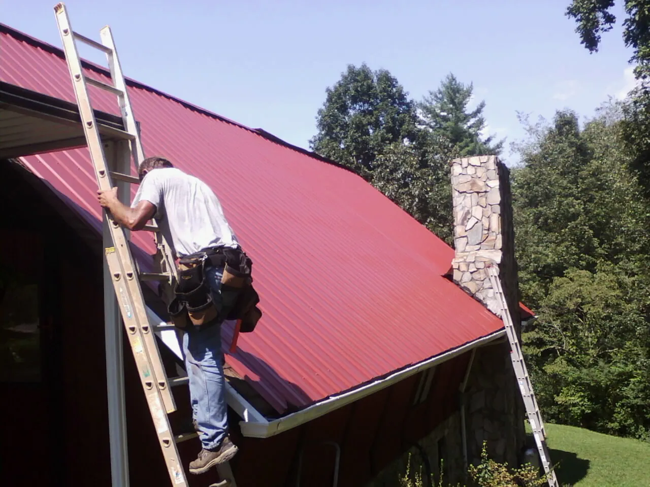 Roofer on a ladder beside a red metal roof and stone chimney — Soffit & Fascia Repair by Roofing Brothers in Huntertown